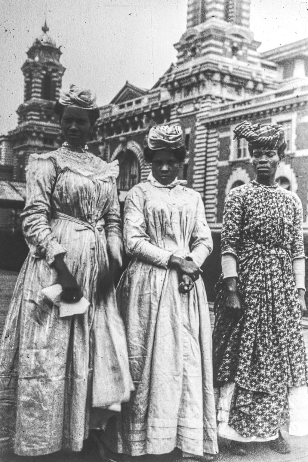 Three women from Guadeloupe photographed at Ellis Island, the main building behind them. Public-domain photograph by Augustus Sherman.